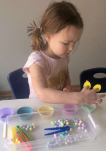 Preschool girl using tweezers to sort colorful objects into muffin cups for fine motor practice