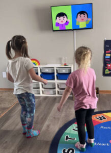 Two preschool girls following movement activities on a TV during indoor learning time
