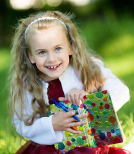 Smiling preschool girl sitting in the grass reading a book with a pen on a sunny day