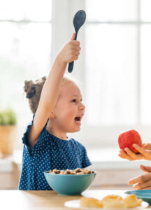 Smiling preschool girl eating cereal and holding a spoon while being offered an apple